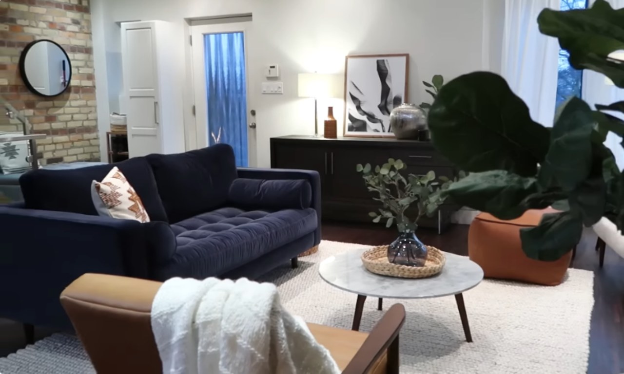 A stylish, modern living room scene featuring a navy blue velvet tufted sofa, a light marble-top coffee table with a woven tray and vase, a tan leather accent chair, and a white rug. The background includes a dark wood credenza with a lamp and abstract framed art, and a brick accent wall on the left.