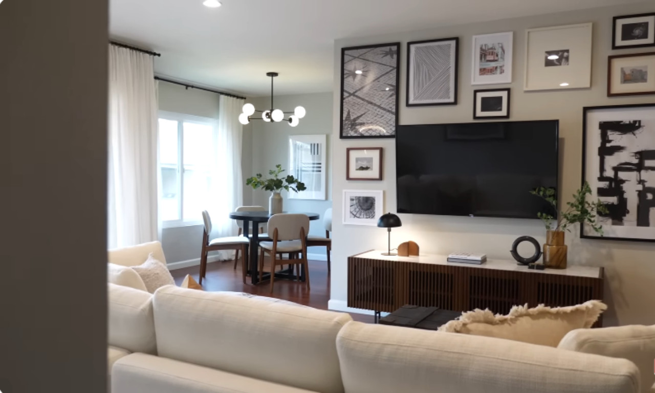 A wide shot of a modern, well-decorated living and dining area. The living room features a white sectional sofa, a console table with a TV mounted above it, and a large gallery wall of framed art. The adjacent dining area has a small round table with four chairs under a contemporary chandelier, with white sheer curtains covering the windows.