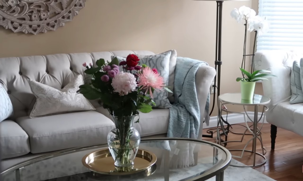 A bouquet of fresh flowers, including pink and red blooms, in a glass vase on a round glass coffee table in a bright, modern living room with a neutral-colored sofa and soft blue and white accents.