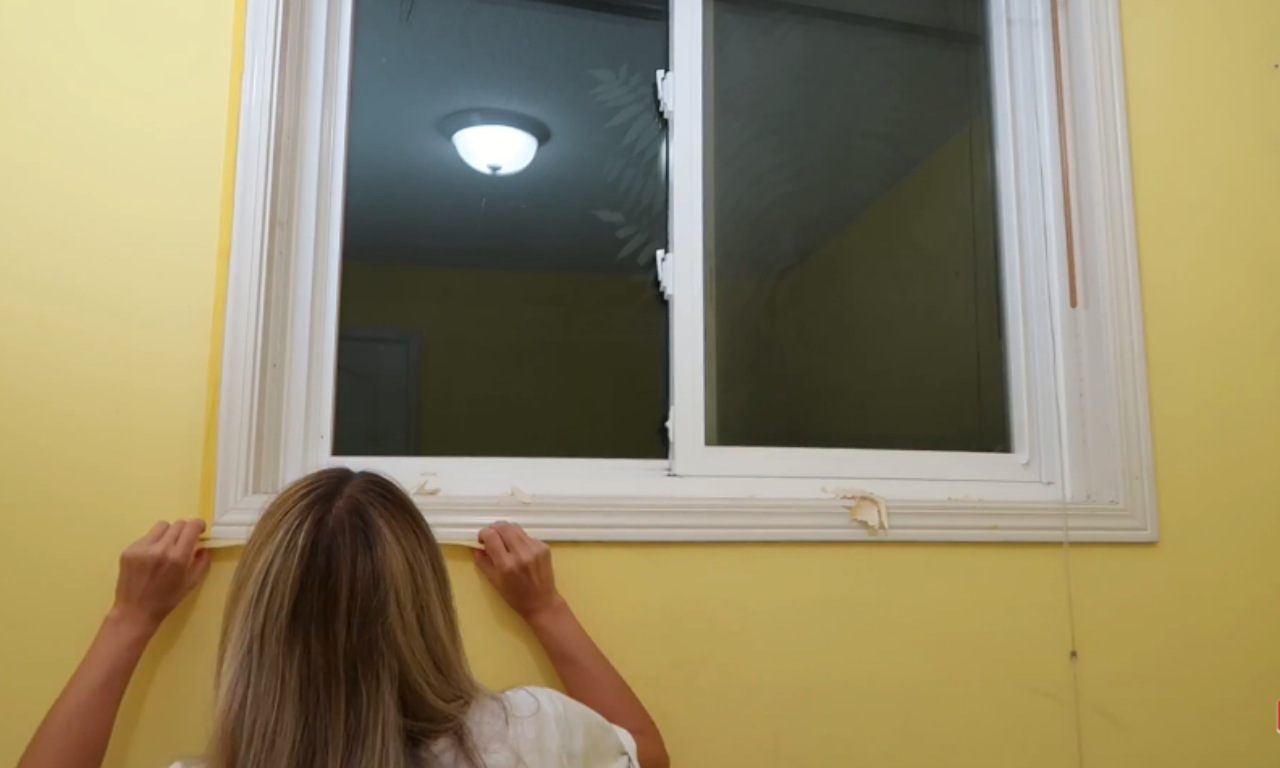 A woman with long blonde hair facing a window, gently pulling a thin strip of material (possibly old caulk or wallpaper trim) from the white trim surrounding the window. The wall around the window is painted bright yellow.