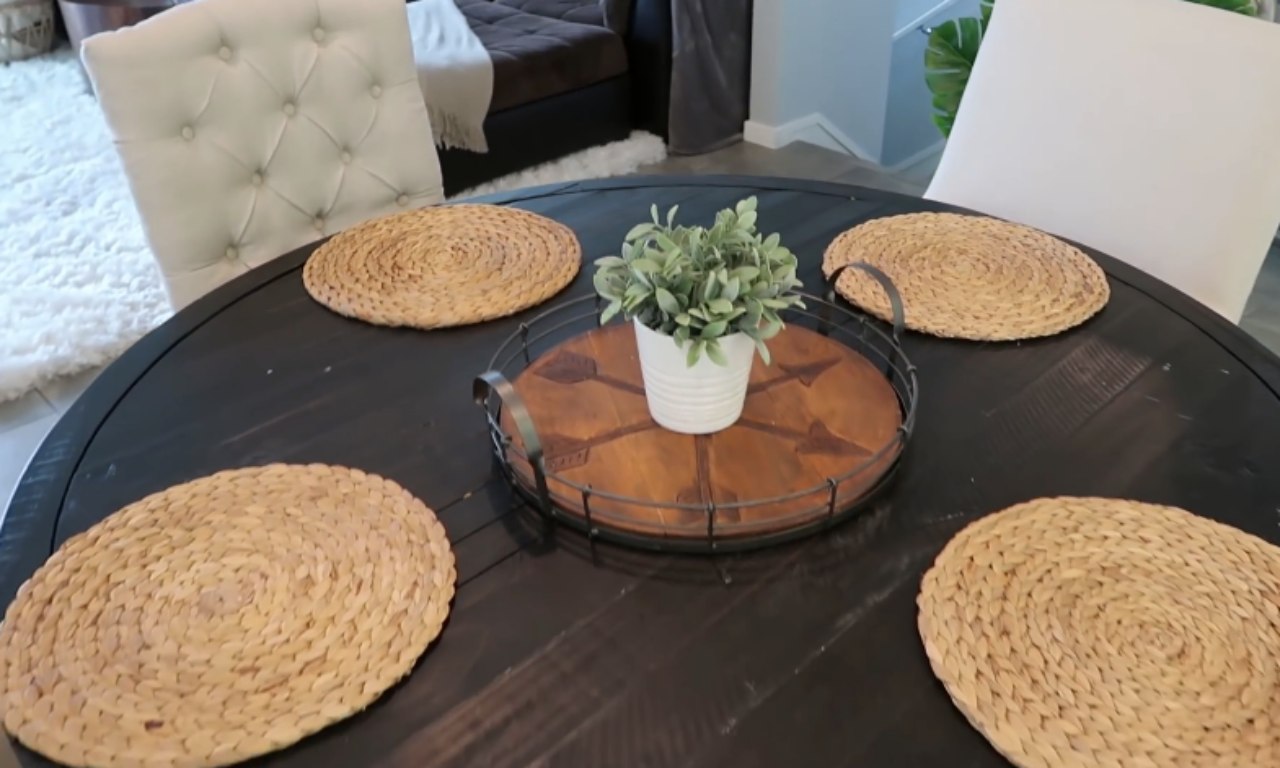 A dark, round wooden dining table set with four woven, circular placemats. A small wooden tray with a metal rim holds a white pot with a green plant as the centerpiece. Parts of two upholstered dining chairs are visible.