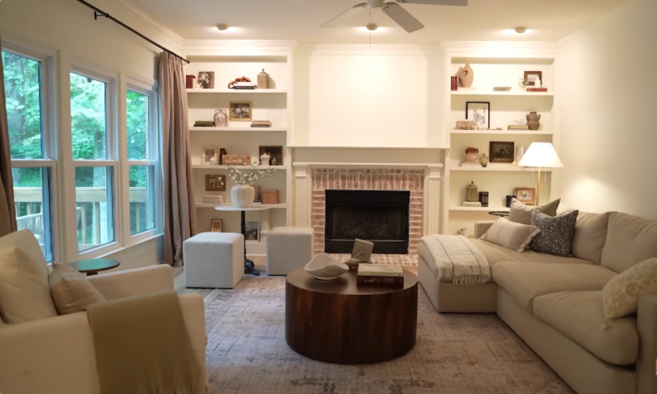 A full view of a bright and inviting living room featuring a neutral color palette. The room has a large sectional sofa, a round wooden coffee table, and a couple of armchairs. The focal point is a brick fireplace flanked by symmetrical white built-in bookshelves, all styled with decorative accents. Large windows on the left look out onto a deck and greenery.