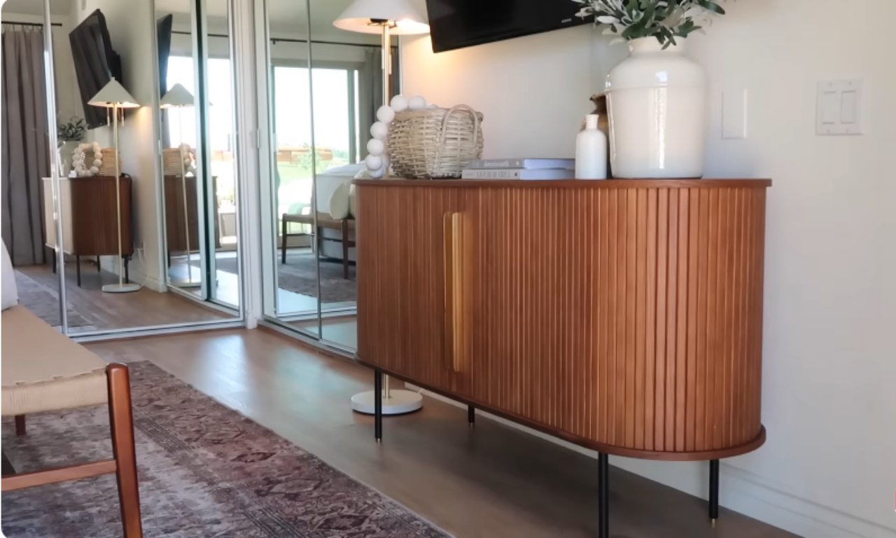 A close-up shot of a modern, mid-century style wooden console or cabinet. The cabinet has rounded sides and a slatted, curved wooden surface in a warm reddish-brown tone, with a single vertical brass handle and thin black metal legs with brass feet. It is set against a white wall next to a wall of mirrored sliding doors. Various decor items, including a white ceramic vase, books, and a woven basket, are placed on top.
