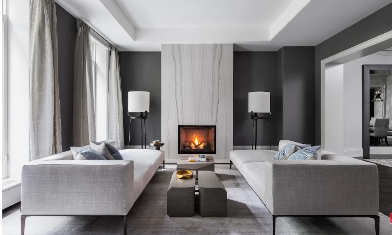 A modern and sophisticated living room with dark gray walls and a white marble fireplace accent wall. Two light gray, minimalist sofas face each other with two slender floor lamps placed symmetrically behind them. A low, dark gray coffee table sits in the center, and the fireplace is lit.