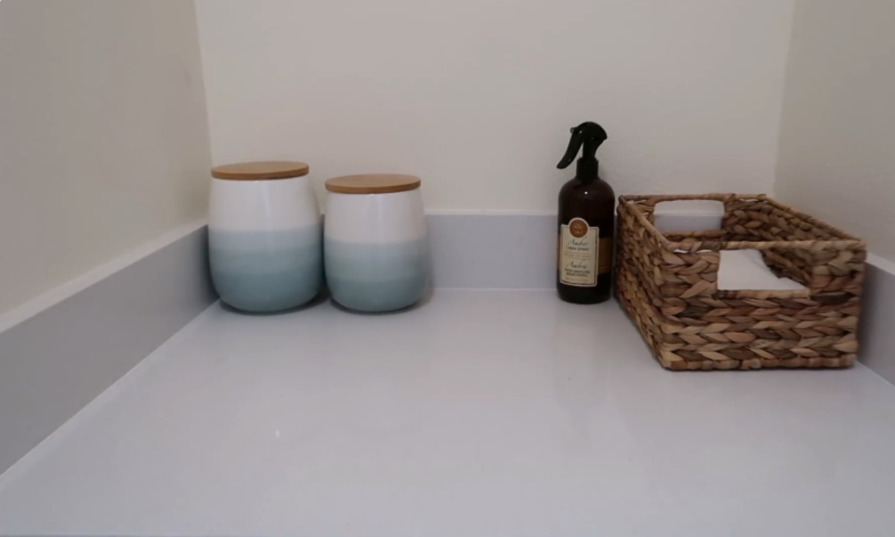 A clean, white countertop is organized with laundry supplies against a white wall. On the counter are two matching ceramic canisters with light blue/aqua gradient bases and wooden lids, a dark spray bottle, and a small woven wicker basket holding various small items.
