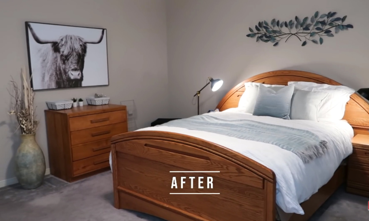 A master bedroom featuring a wooden bed frame with white and light blue bedding, a bedside lamp, and a tall wooden dresser with decorative baskets. A large, framed black-and-white print of a Highland cow hangs above the dresser, and a tall vase with dried plants is next to it.