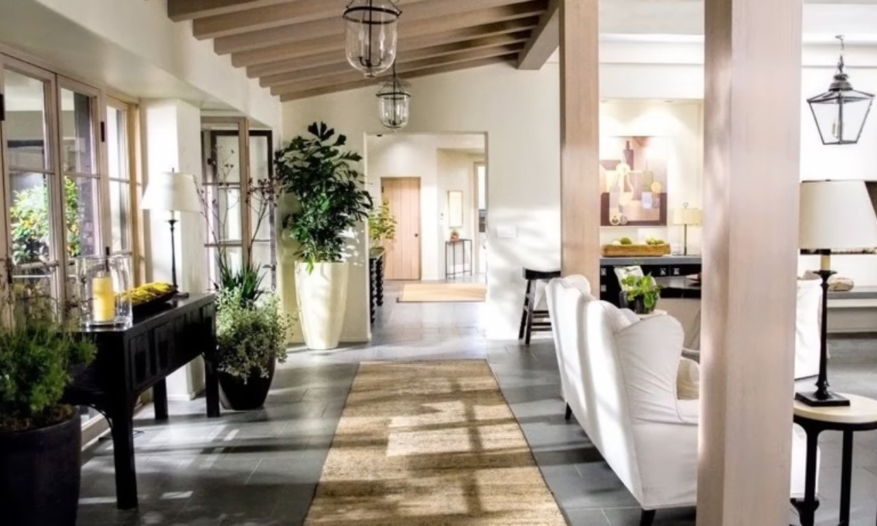 A bright, spacious entryway or hallway with a vaulted wooden ceiling, stone tile floor, and a light-colored rug running down the center. Tall planters with green plants and black console tables are visible, along with white slipcovered furniture in the distance.