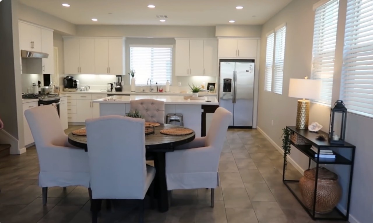 A view of a modern open-plan kitchen and dining area. The kitchen features white cabinets, a stainless steel refrigerator, and a center island. In the foreground, there is a round dark wood dining table surrounded by four light-colored upholstered chairs. To the right, a narrow black console table is styled with a lamp, a lantern, and a decorative woven basket on the floor beneath it.