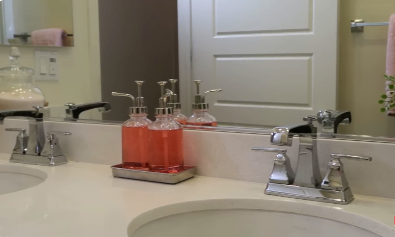 A close-up view of a double vanity in a modern bathroom. The countertop is white, and the chrome faucets are polished. In the center, there is a silver tray holding three clear glass soap/lotion dispensers filled with a coral-pink liquid. A large mirror runs the length of the vanity, reflecting the dispensers and the opposite white door.