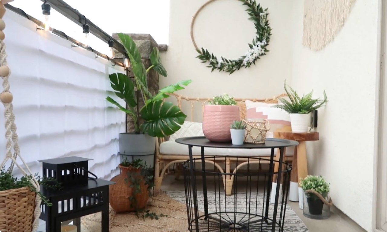 A view of a small, renter-friendly balcony makeover featuring a woven rattan loveseat with pillows, several potted faux plants (including a large Monstera), and a round, black metal basket-style coffee table. The balcony railing is covered with a white sheet or privacy screen, and fairy lights are strung above. A simple green wreath hangs on the back wall.