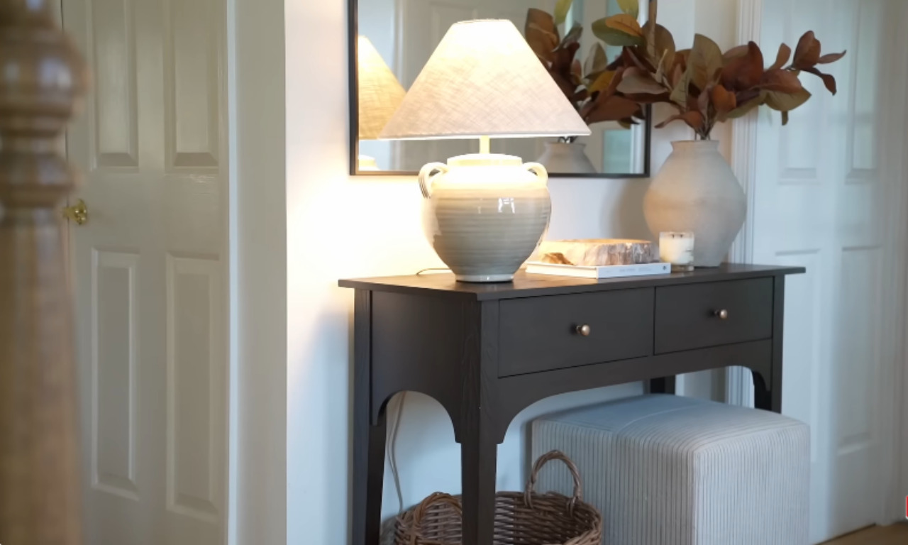 A dark wood console table styled with a beige ceramic table lamp, a rectangular mirror above it, and a large, textured vase holding brown leaves. A striped ottoman is tucked underneath, and a woven basket rests on the floor beside it in a well-lit hallway.
