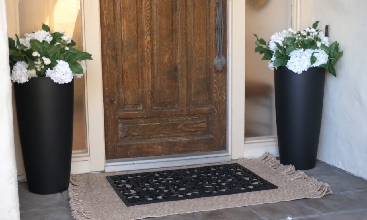 An exterior shot of a wooden front door with a glass side panel, flanked by two tall, black planters containing large white hydrangea arrangements. A natural fiber rug with a black, patterned doormat is placed in front of the door.