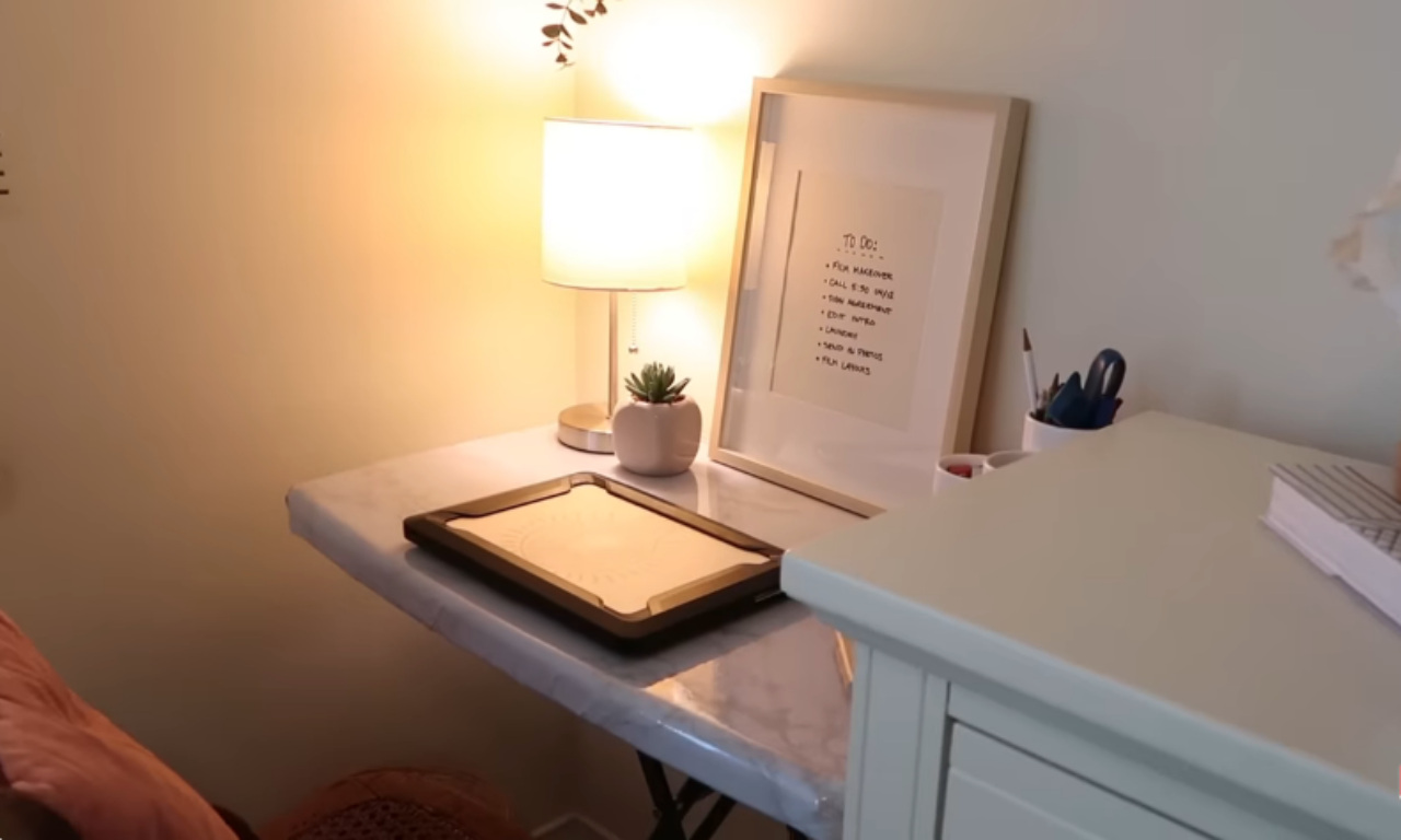 A wider view of a brightly lit, small home office corner. The desk has a marble-look surface. On it are a silver table lamp, a framed handwritten "TO DO" list, a potted succulent, and a black digital notepad in a tray. To the right is a white cabinet or drawer unit, creating an L-shape with the desk. The overall look is clean and minimalist.