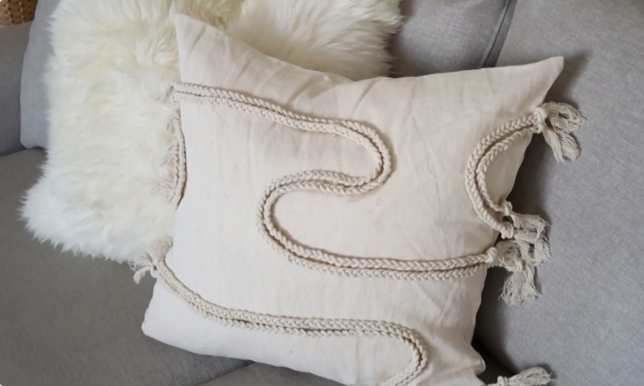 A close-up of a cream-colored throw pillow on a gray couch, decorated with a swirling, braided beige cord and tassels on the side, next to a fluffy white sheepskin throw.