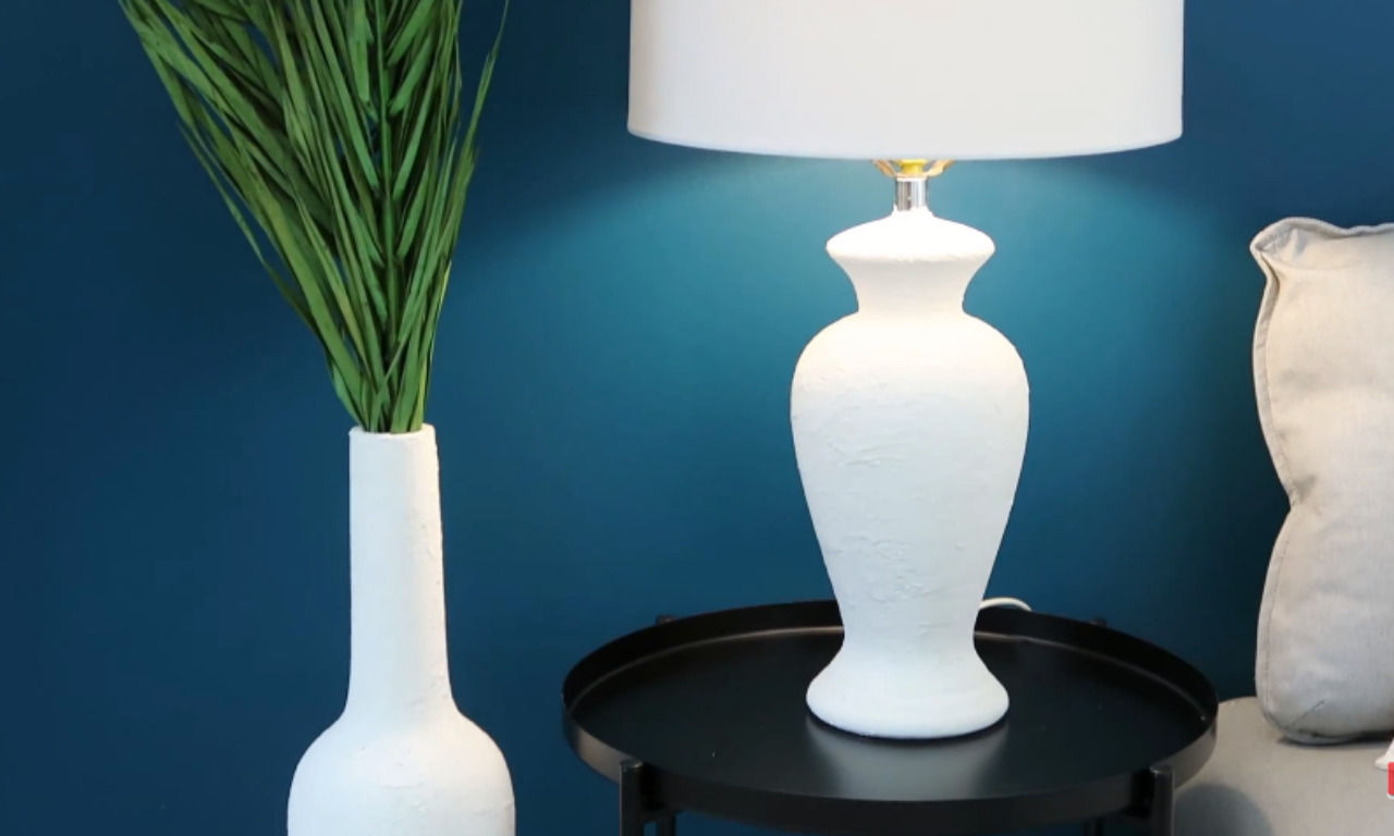 A textured white ceramic lamp with a white lampshade on a small black side table, next to a matching white ceramic vase holding tall green decorative grasses, set against a deep teal blue wall.
