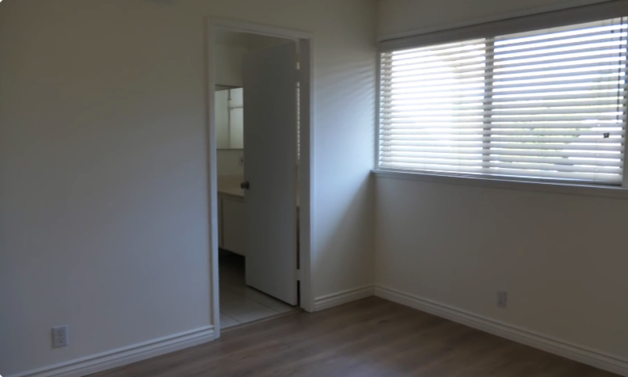 An empty, sunlit bedroom with light wood flooring and plain white walls. The room features a window with white horizontal blinds and an open doorway leading into an en suite bathroom with a vanity visible inside.