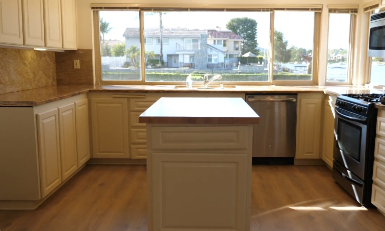A bright, empty kitchen with white-painted cabinets, light brown granite countertops, and light wood flooring. A window over the sink looks out onto a canal and houses. A small central island with a butcher block top is in the middle of the room. Appliances include a dishwasher and a black stove.