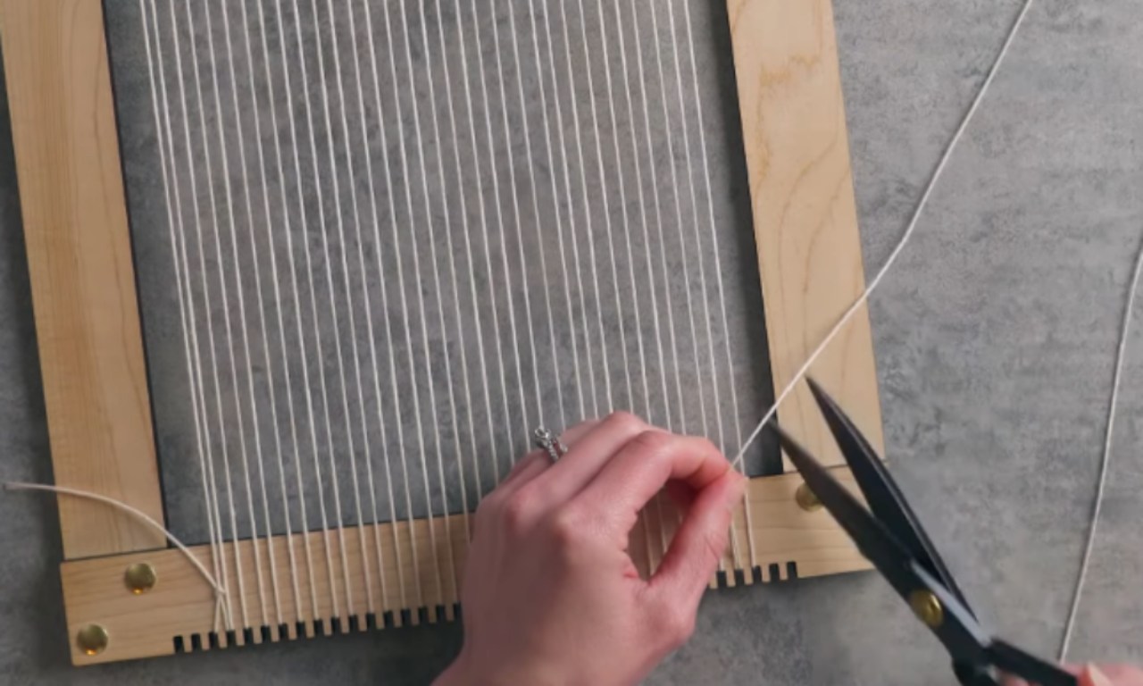 A person cuts the white warp string with black scissors, finishing the process of warping a wooden frame loom that is filled with vertical white threads.
