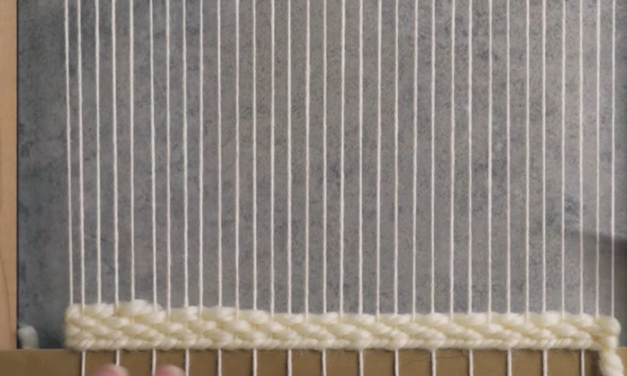A close-up of a frame loom showing the foundation of a weaving project. Thick, cream-colored yarn is woven in a textured pattern for the first few rows at the bottom of the vertical white warp threads.