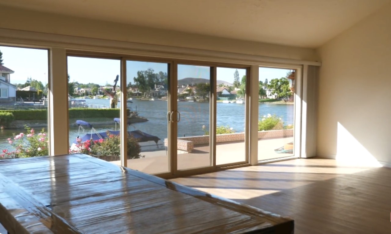 An empty living or dining room with light wood floors. The room features a large, glass sliding door that opens to a patio, a lake, and houses on the opposite bank. A large, plastic-wrapped rectangular object, possibly a dining table, sits in the foreground.