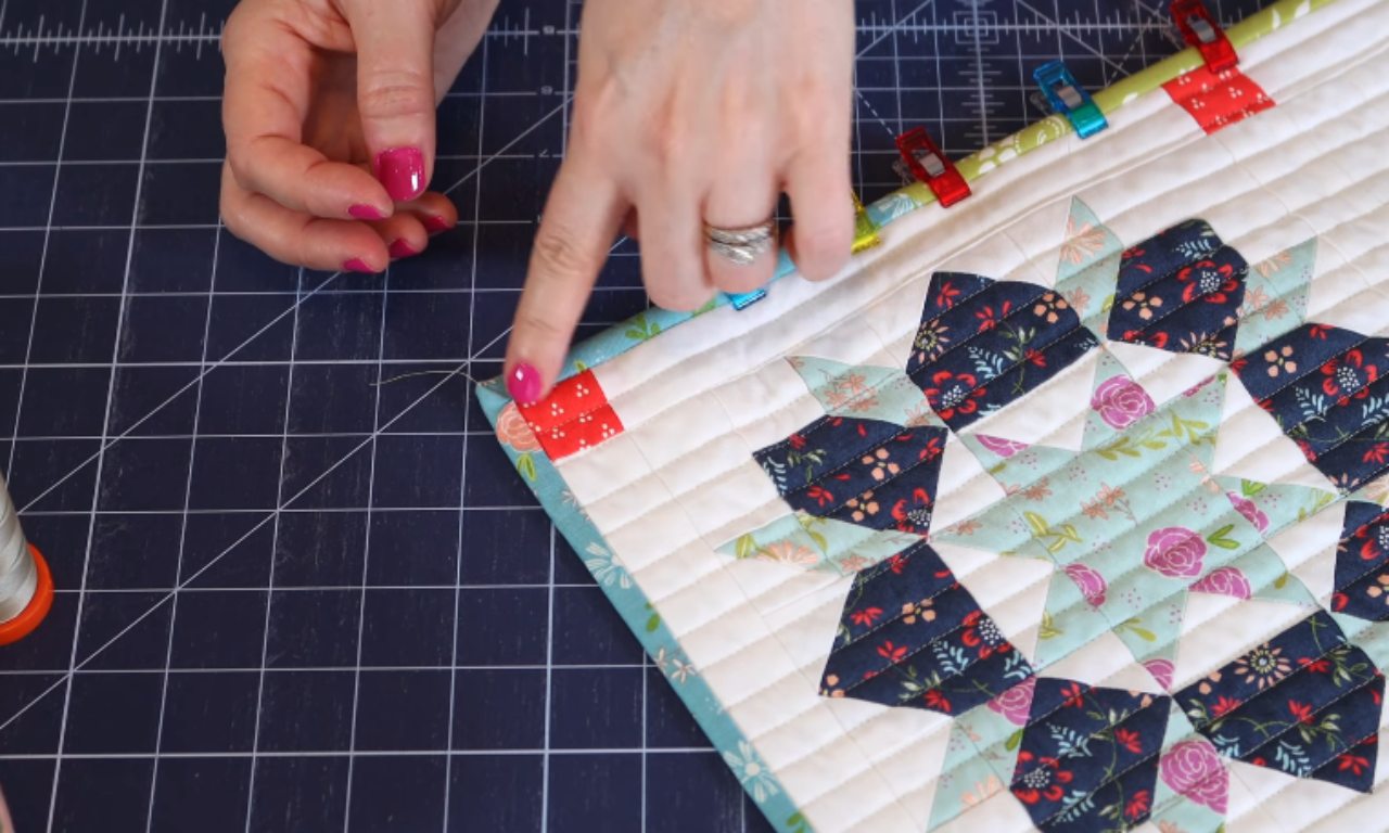 A close-up of a quilter demonstrating how to hand stitch a mitered corner on a floral quilt, pointing to where the thread secures the binding.