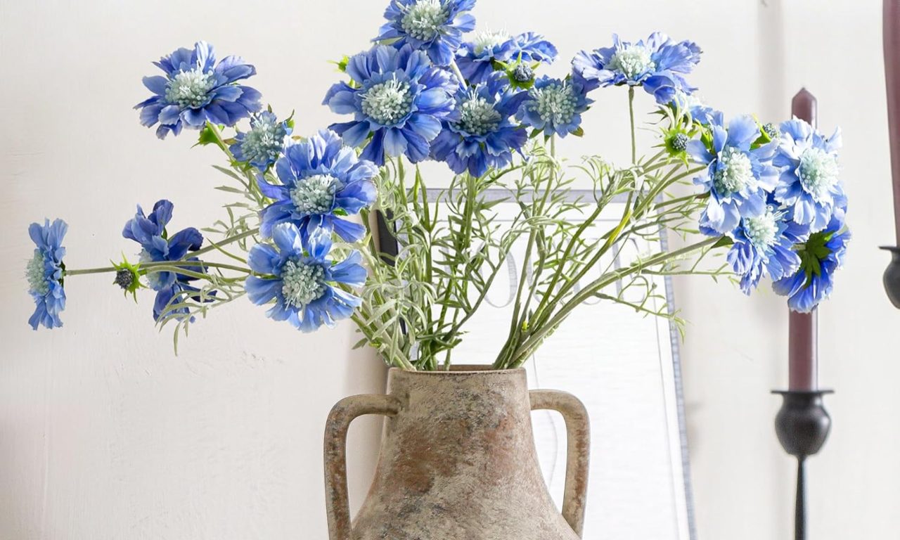 Close-up of bright blue cornflowers arranged in a rustic, earth-toned Mitt&Ditt ceramic flower vase. The scene features a farmhouse decor style with a soft-focus white background and a dark candle holder, highlighting the textured pottery and vibrant floral colors.