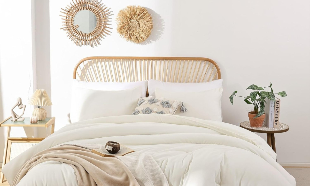 A cozy bohemian style bedroom features a wooden spindle headboard bed with soft white comforter and pillows, a patterned throw pillow, two bedside tables with lamps and a potted plant, and a sunburst rattan mirror and matching straw decor on the wall.