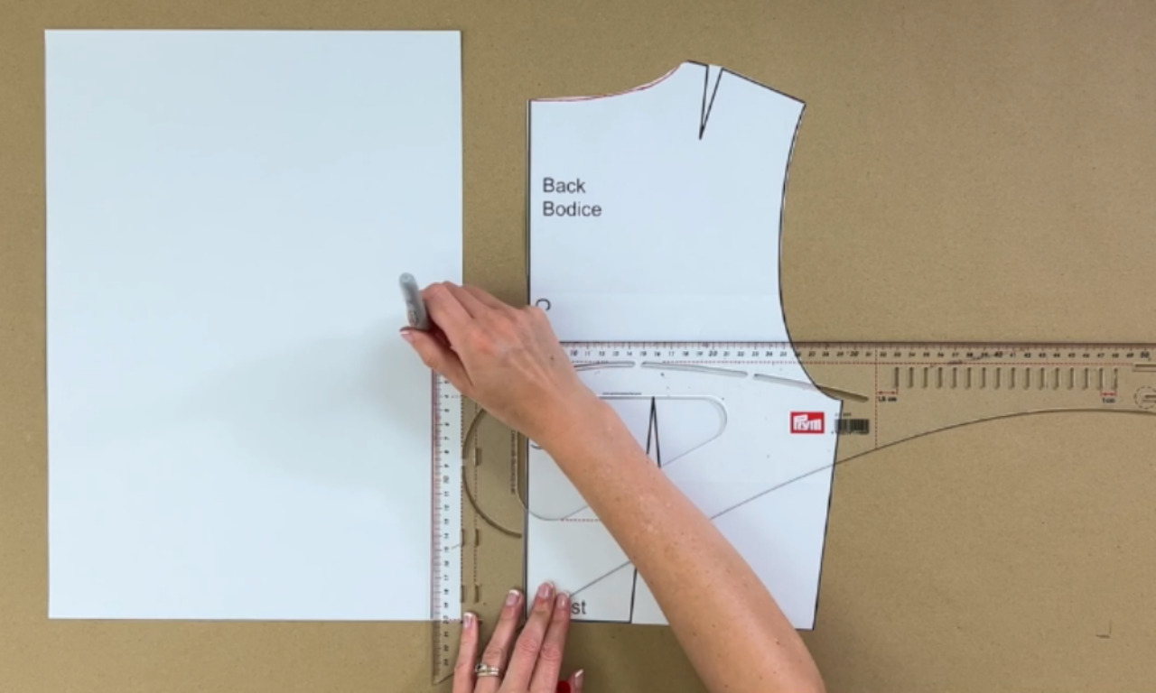 A person using a clear pattern master ruler to draft a line next to a back bodice paper pattern.
