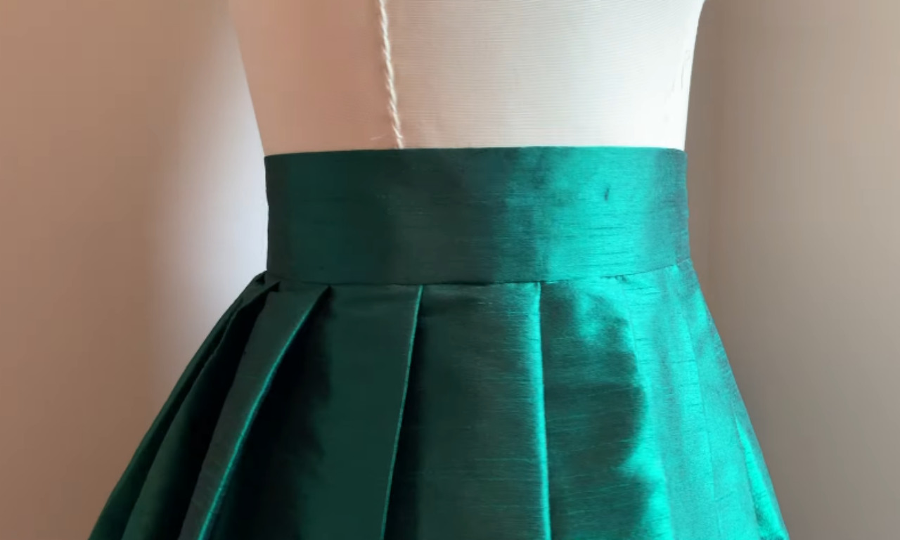 A close-up view of the final professionally sewn waistband on a green pleated skirt, displayed on a dress form.