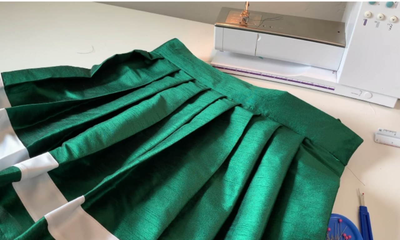 A shiny green pleated skirt with a newly attached waistband sits on a white worktable with a sewing machine in the background.