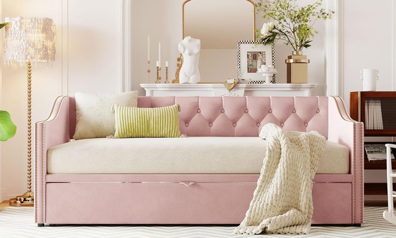 A velvet pink daybed with a tufted backrest and a pop-up trundle, designed to convert from a twin to a king size for small space multi-use guest rooms. The bed is styled with light-colored pillows and a throw blanket in a modern, bright living area.