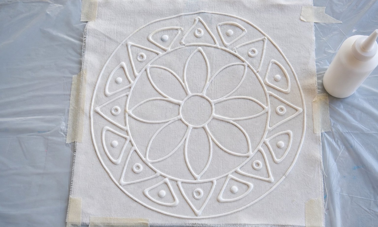 Thick white glue lines traced over a pencil leaf design on white cotton, with the glue bottle nozzle resting beside the fabric on a protected table