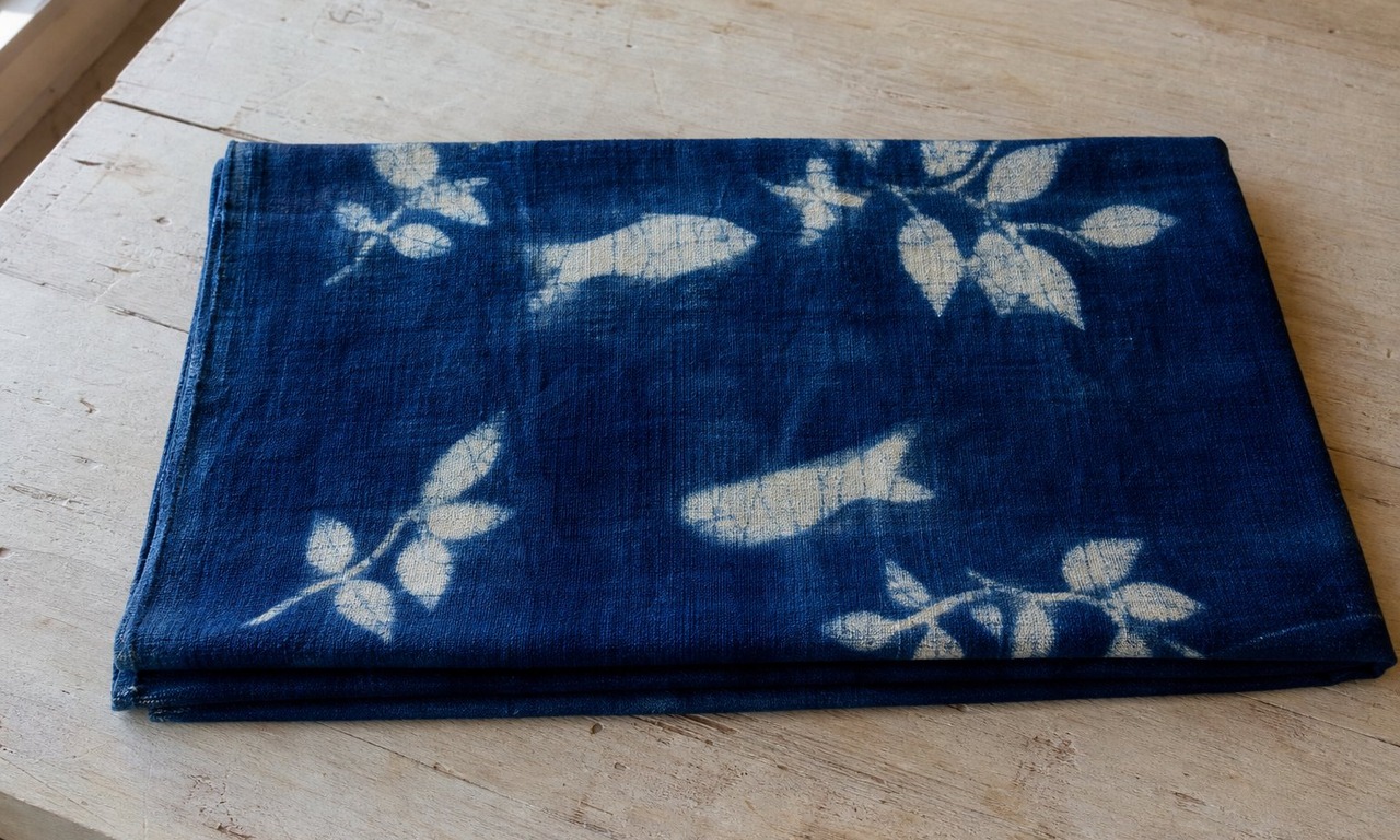 A folded length of indigo dyed cotton with pale leaf shapes standing out against the darker ground, photographed flat on a pale wood surface