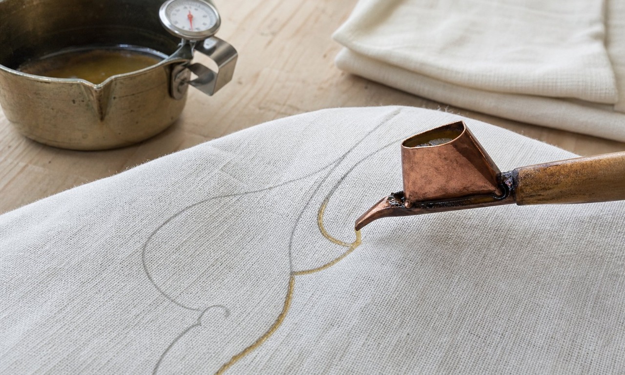 A small copper canting tool drawing a fine wax line along a pencil curve on white cotton, with a wax pot and thermometer behind on a pale work table