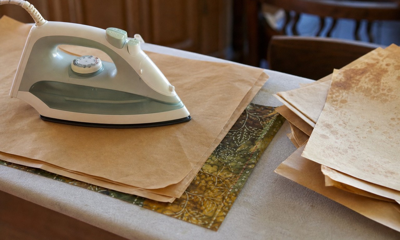 A warm iron pressing a folded batik panel between layers of brown paper on an ironing board, with used paper showing faint wax marks to one side