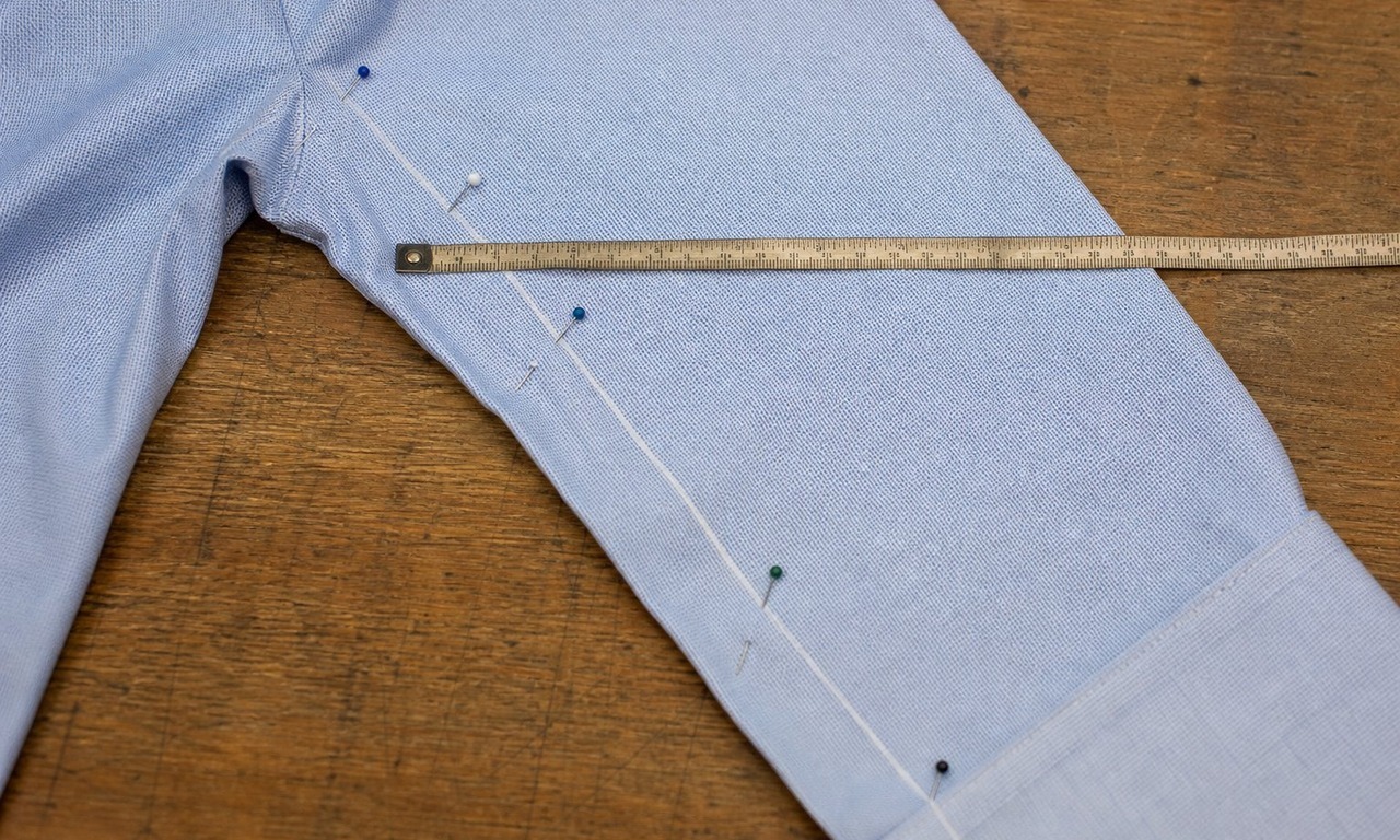 A chalk line tapering down the underarm sleeve seam of a blue oxford shirt, with a tape measure across the bicep